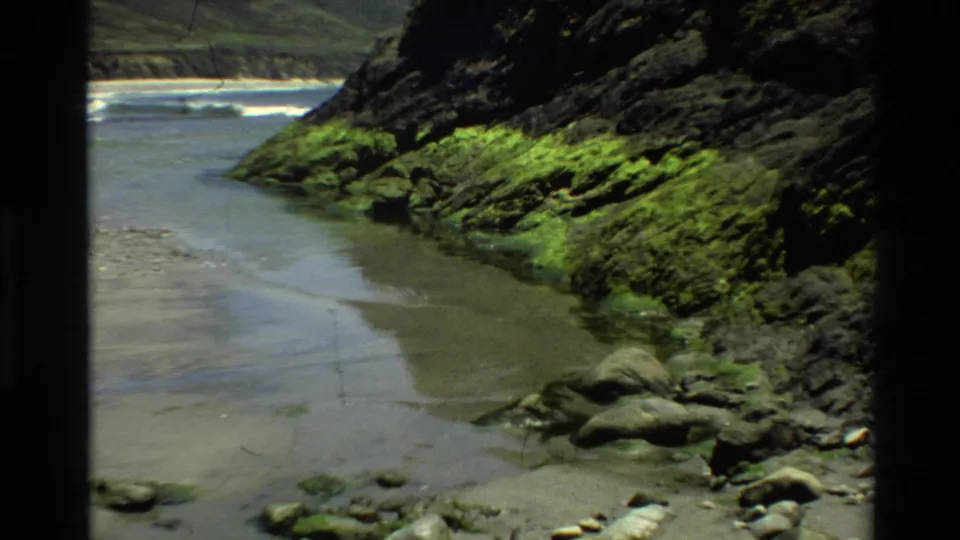 Super 8 close coastal shot likely from Big Sur circa 1980, showing a moss-covered rocky cliff, narrow tidal cove, and rounded boulders.