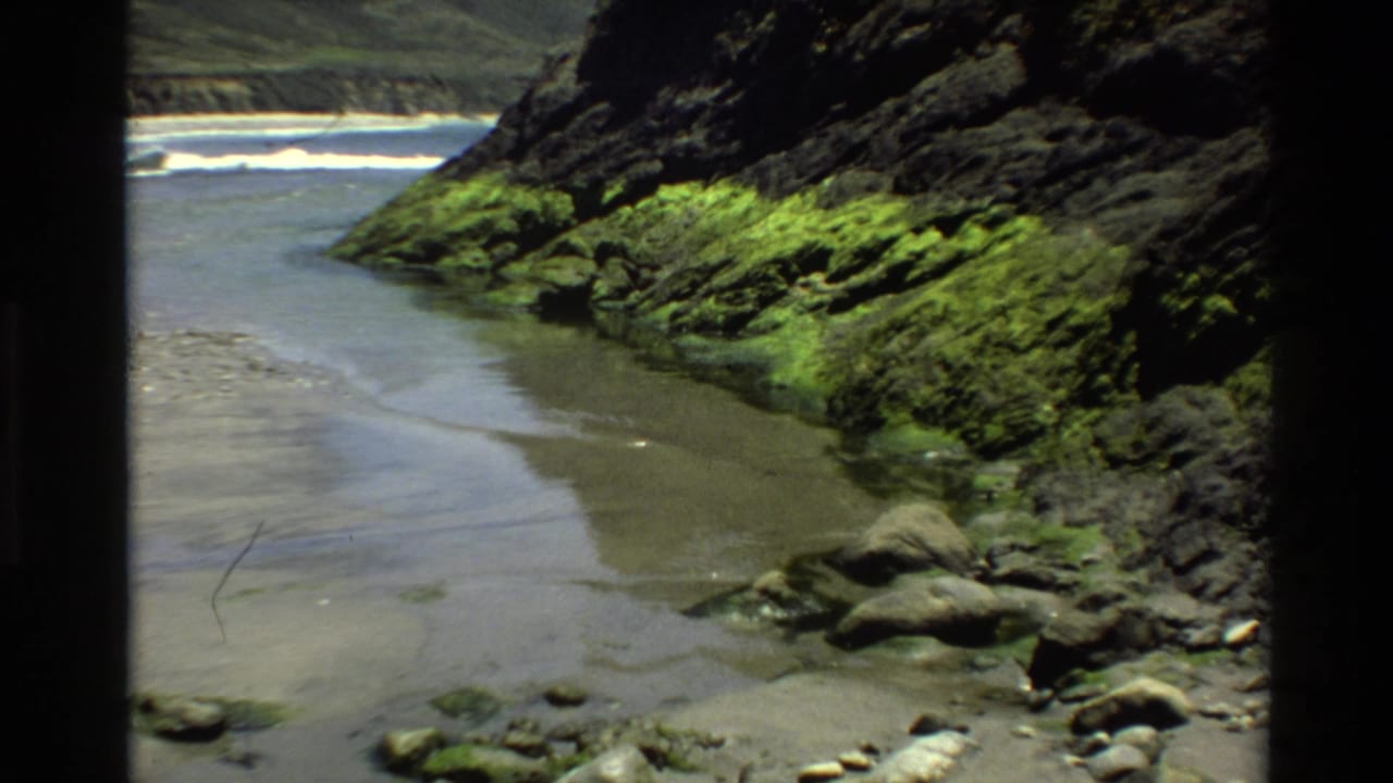 8mm home movie frame from circa 1980 showing a sandy tidal inlet at Big Sur, California, flanked by moss-covered volcanic rock cliffs, with surf visible at the cove mouth.