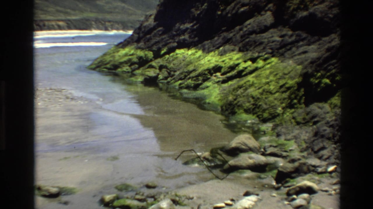 8mm footage from circa 1980 showing the same Big Sur coastal inlet, with green moss-draped volcanic rocks framing a sandy tidal channel and distant whitecaps.