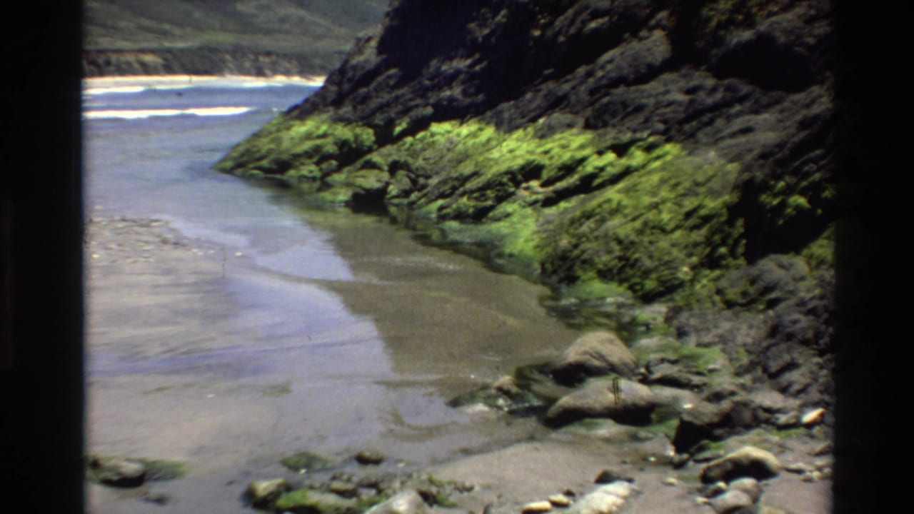 Mid-clip 8mm frame from circa 1980 showing the Big Sur coastal inlet; moss-covered volcanic rock cliffs and a shallow sandy channel remain the central subject.