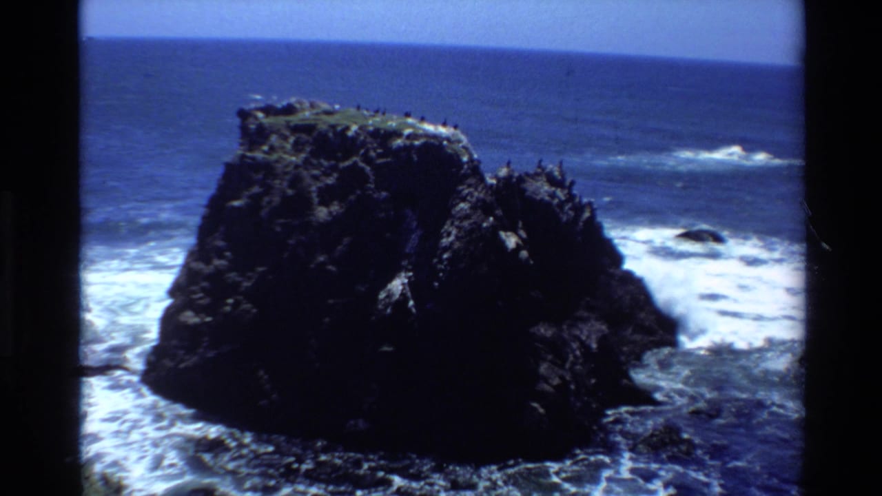 1980s 8mm home movie frame showing a large dark coastal rock with seabirds, white surf at its base, and blue Pacific Ocean, likely Big Sur, California.