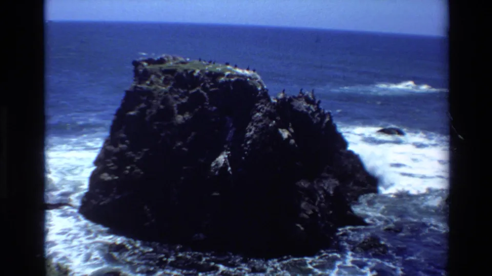 1980s 8mm home movie frame showing a large dark coastal rock with seabirds, white surf at its base, and blue Pacific Ocean, likely Big Sur, California.