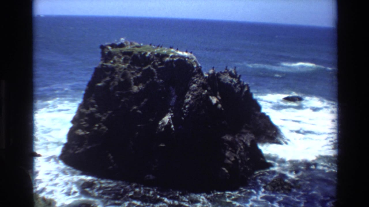 Large dark coastal rock formation with seabirds on top, white surf and spray at the base, blue sky and open ocean.