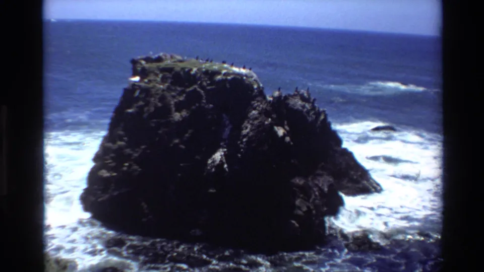 1980s 8mm home movie frame of a rugged coastal rock with seabirds, breaking Pacific waves, and clear blue sky, likely Big Sur, California.