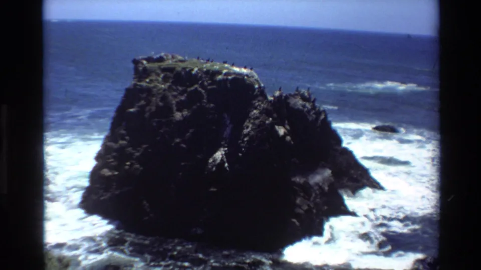 Closing 8mm frame of a coastal sea stack with roosting seabirds and Pacific surf, likely Big Sur, California, dated to 1980.