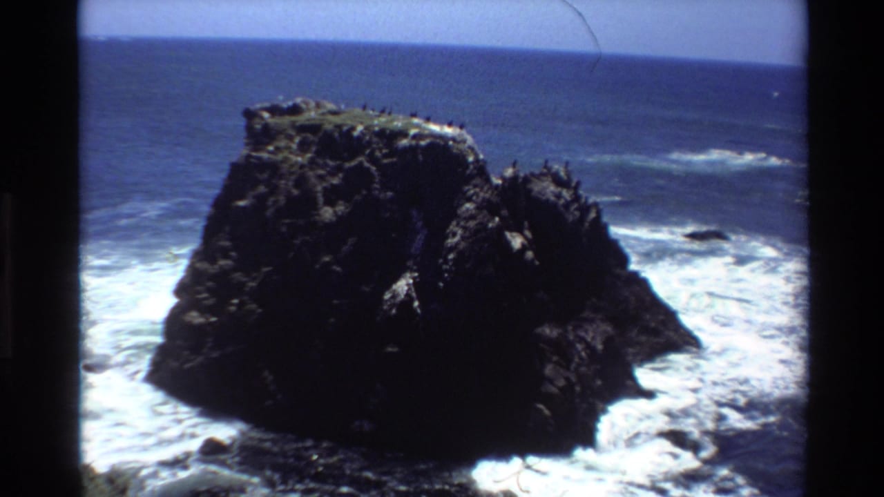 Super 8 frame of a dark basalt sea stack off the California coast circa 1980, with breaking surf and seabirds roosting at the top.