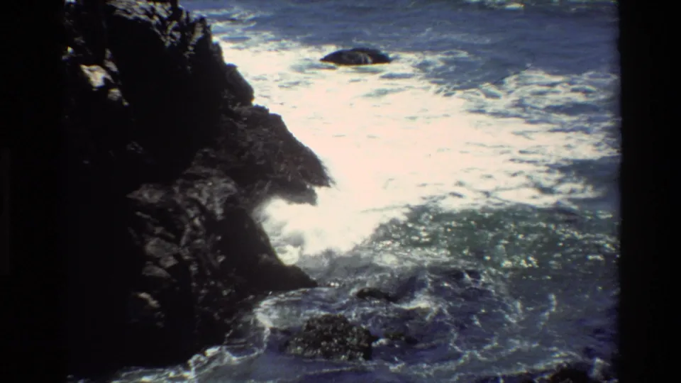 Rocky Pacific shore, circa 1980, likely Big Sur — 8mm home movie frame showing waves breaking at the base of a dark coastal rock.