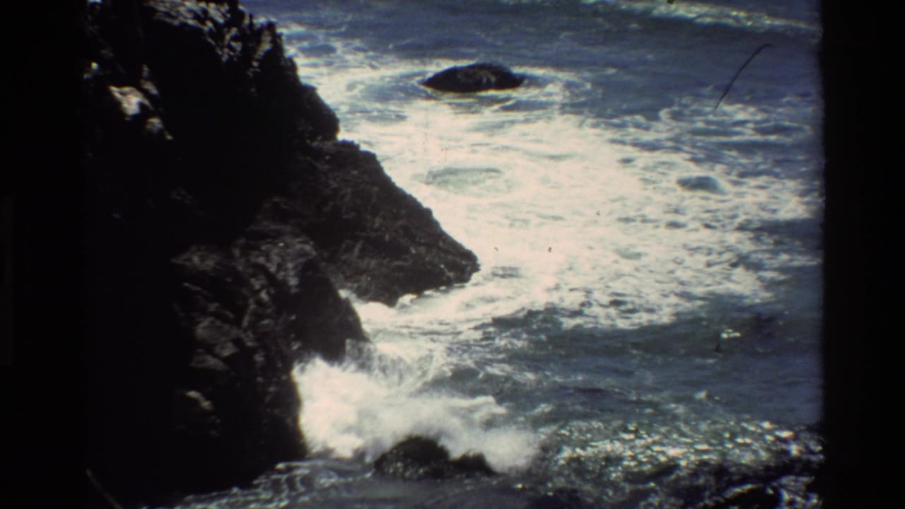 1980s 8mm film frame showing dark Pacific coastal rocks with white surf crashing, Big Sur, California.