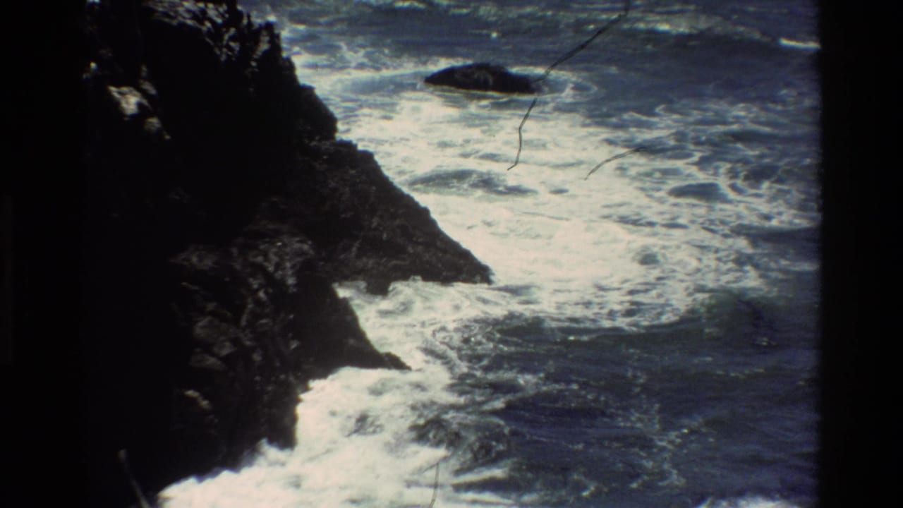 1980s 8mm home-movie frame of Pacific surf breaking on dark rocks, Big Sur coastline, California.