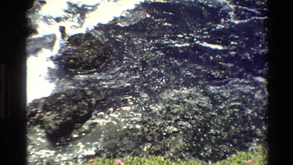 1980s 8mm film frame showing glistening dark rocks and white surf foam at the Pacific coastline, California.