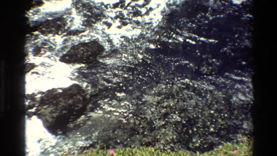 1980s 8mm home-movie frame of coastal rocks and churning Pacific surf with grass at foreground edge, California coast.