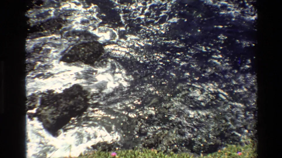 Super 8 home movie, c. 1980, Big Sur, California — water over dark rocks, grassy foreground with small pink markings, possibly wildflowers.