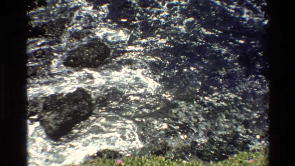 Super 8 home movie, c. 1980, Big Sur, California — sunlit water over wet rocks, grassy lower foreground with small pink markings and characteristic 8mm grain.