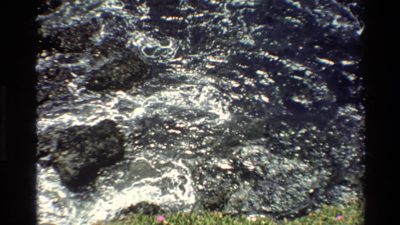 Super 8 close-up of creek water rushing over dark rocks with white foam and green vegetation at the edge, circa 1980, Big Sur, California.