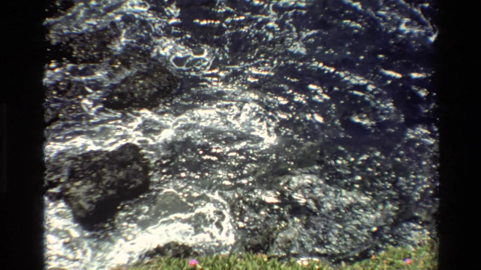 Super 8 close-up of creek water rushing over dark rocks with white foam and green vegetation at the edge, circa 1980, Big Sur, California.