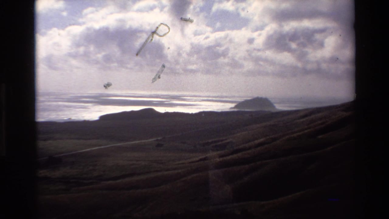 Coastal panorama showing grassy hillside, distant rocky sea formation, ocean horizon, and partly cloudy sky with film emulsion artifacts.