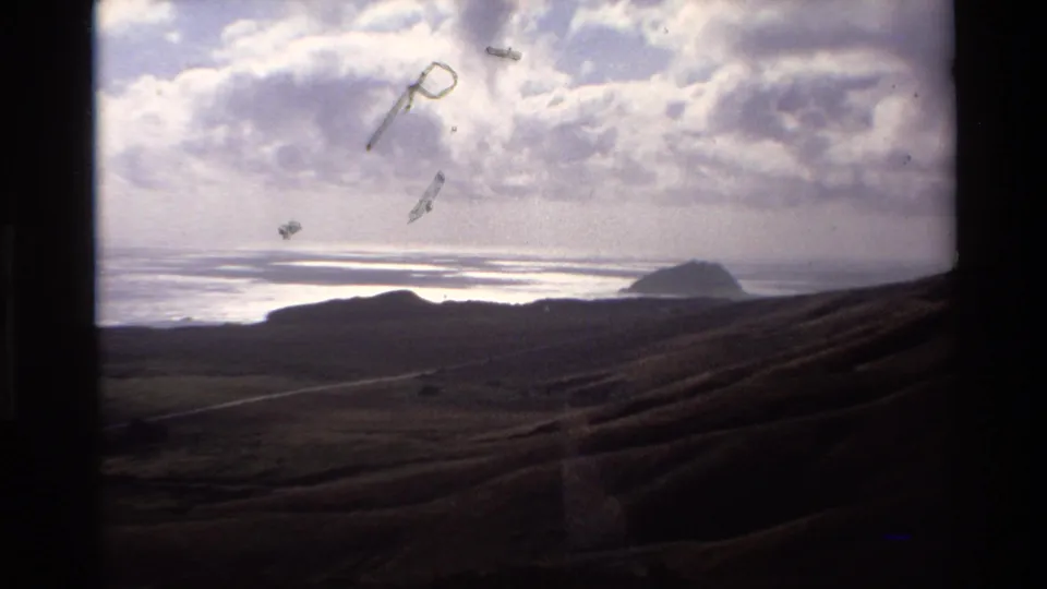Super 8 coastal panorama with rolling hillside, distant sea rock, and overcast sky; emulsion flare artifacts are visible in the upper frame; circa 1980, Big Sur area, California.