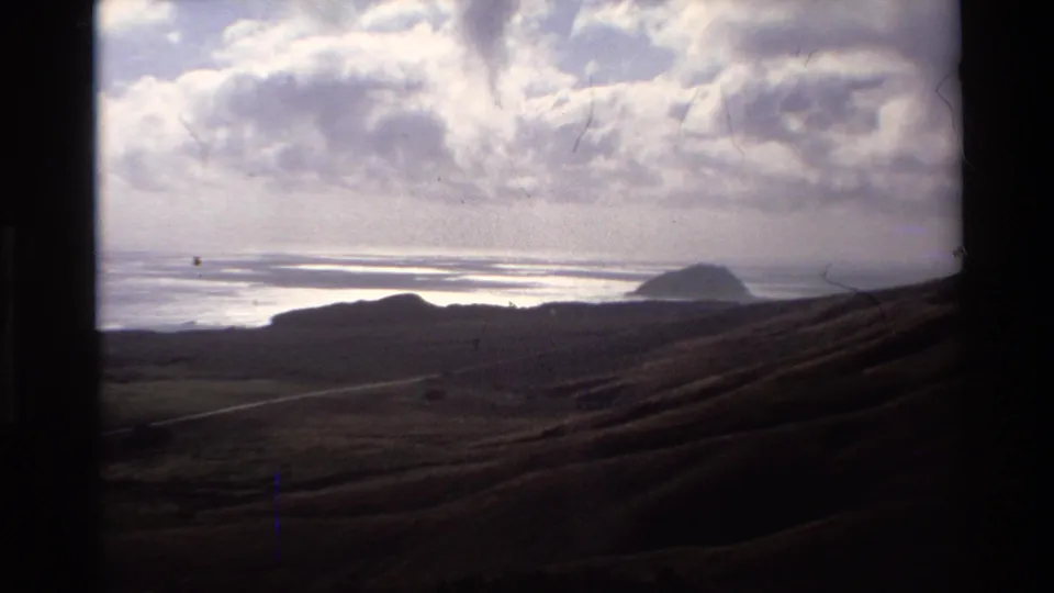 Super 8 wide shot of California coastal hills with a prominent distant sea rock and overcast cloud bank, circa 1980, likely Big Sur area.
