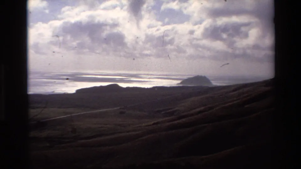 Super 8 coastal panorama with rolling hills and distant sea rock under cloudy sky; hairline film scratches visible across the frame; circa 1980, Big Sur area, California.