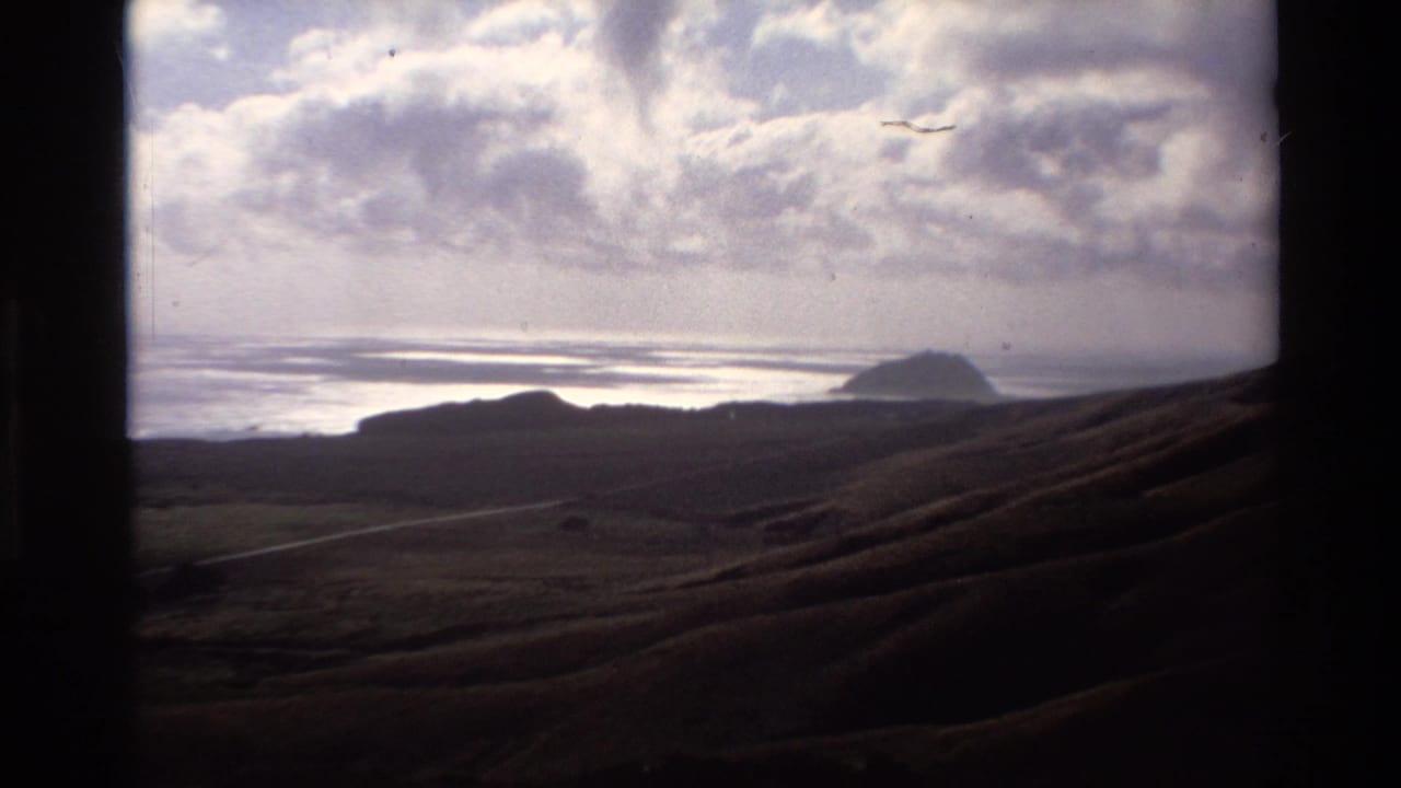 1980s Super 8 home movie of California coast — shadowed coastal hills, calm ocean bay with glinting water, distant headland, and cumulus clouds from an elevated vantage.