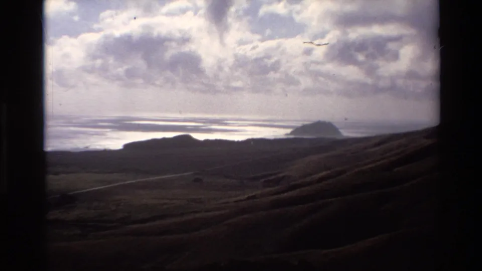 1980s Super 8 home movie of California coast — shadowed coastal hills, calm ocean bay with glinting water, distant headland, and cumulus clouds from an elevated vantage.