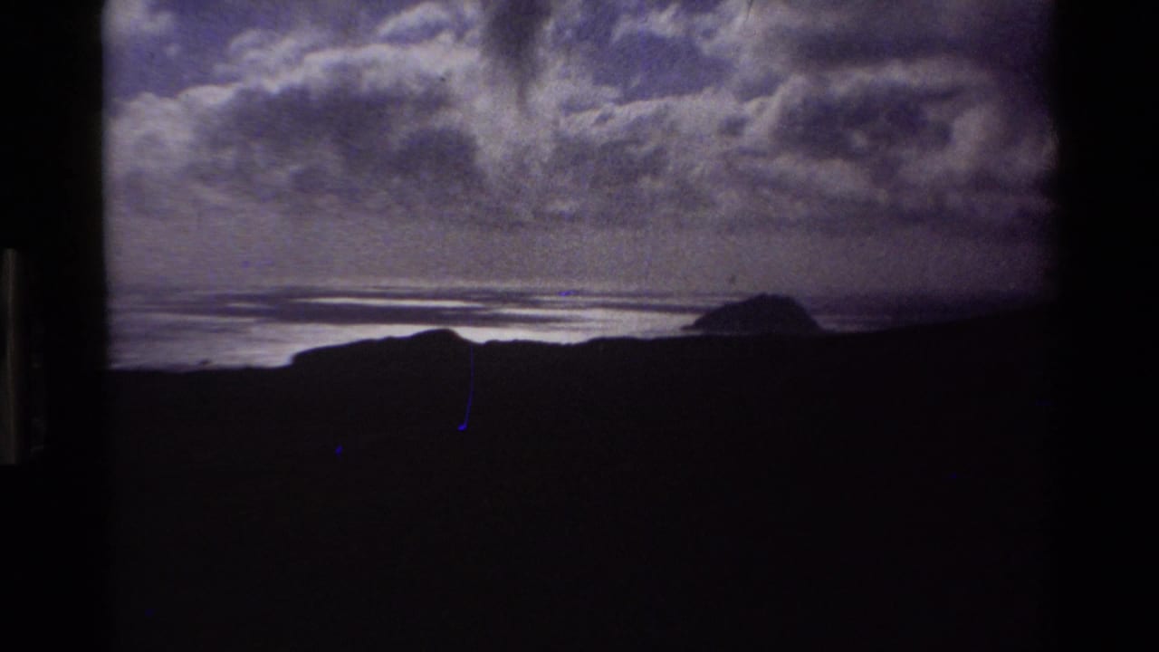 Slightly darker elevated coastal view showing shadowed hills, ocean bay with reflected light, distant headland, and dense cumulus cloud cover on Super 8 film.