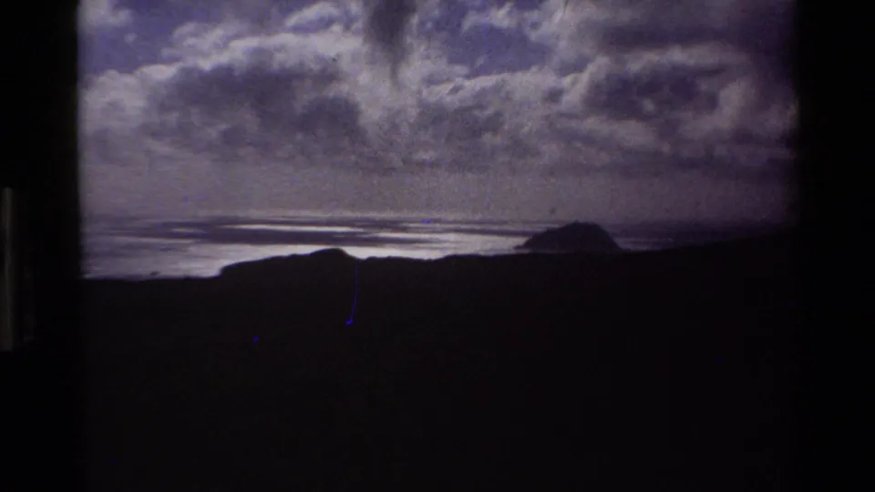 1980s Super 8 frame of California coast in darker exposure — shadowed hills, light-reflecting bay, distant offshore headland, dense cumulus clouds, and a faint vertical film artifact.