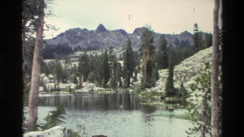 8mm footage circa 1980 of a calm alpine lake framed by conifers and jagged rocky peaks; attributed to the California area per reel metadata.