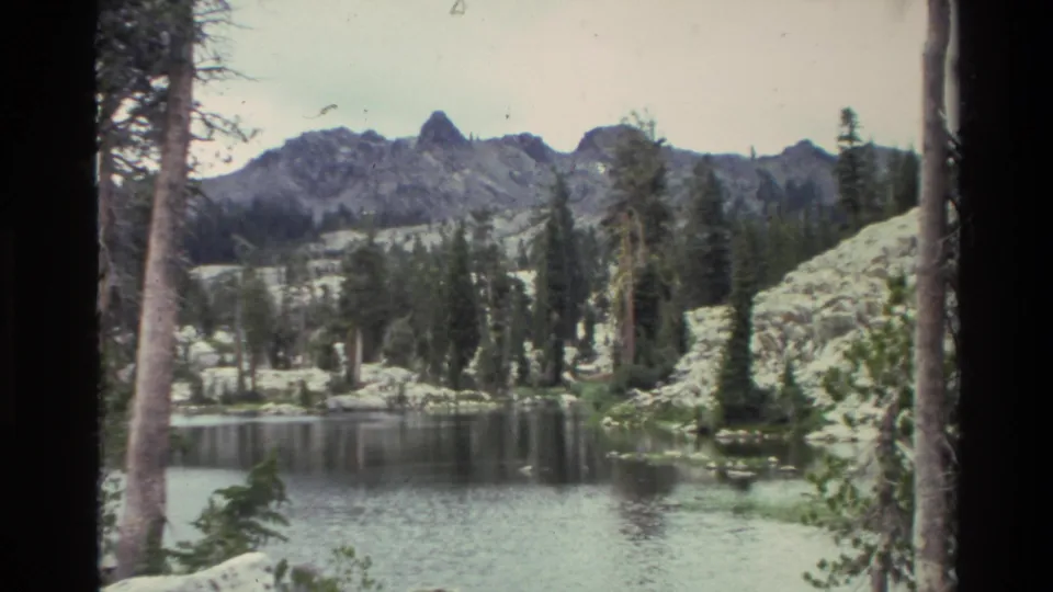 8mm home-movie frame circa 1980 showing a wide alpine lake panorama with conifers, exposed rocky hillside, and rugged peaks in background.