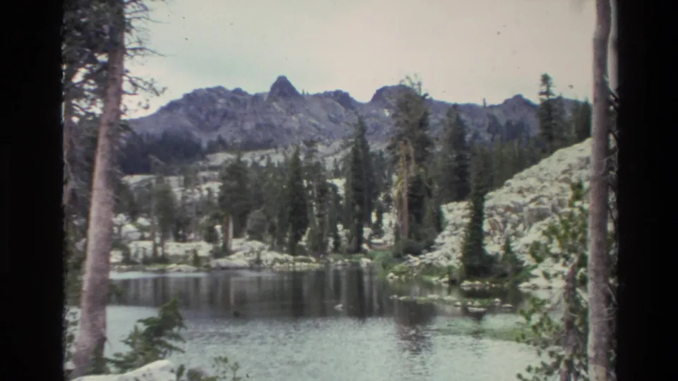 8mm footage circa 1980, near-static composition of an alpine lake with conifer shoreline and rocky peaks; right foreground tree trunk visible.
