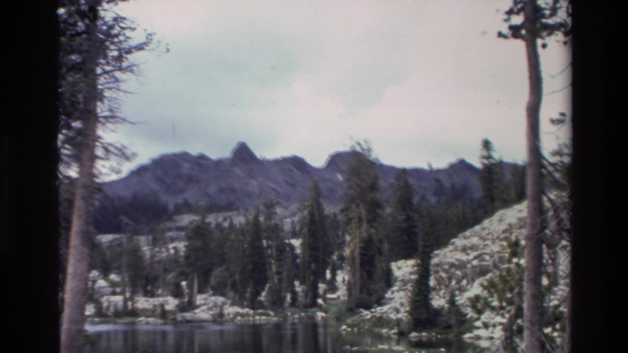 Mountain lake view with conifer-lined rocky shores and rugged peaks, slight camera pan, 8mm grain and colour fading.