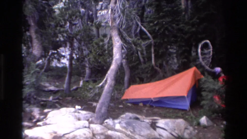 Campsite with orange and blue backpacking tent and partial figure of a person at right edge, gnarled trees and rocky ground, California circa 1980.