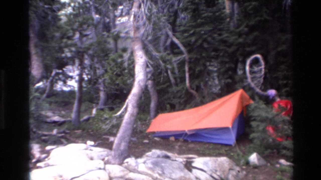 8mm home movie frame: orange-blue dome tent among granite boulders and conifers, figure in red near tent, California forest campsite, c.1980.