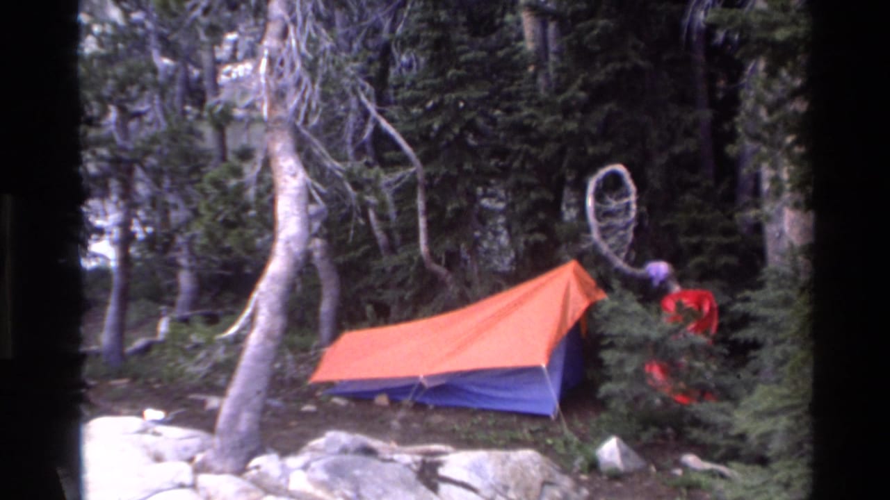Orange and blue tent in rocky conifer forest, figure in red clothing beside tent, bare trees and boulders surrounding the campsite.