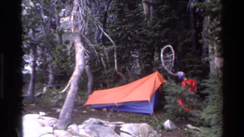 8mm frame of orange-blue dome tent in a California conifer forest, c.1980; figure in red and a circular hoop hanging from a branch also visible.