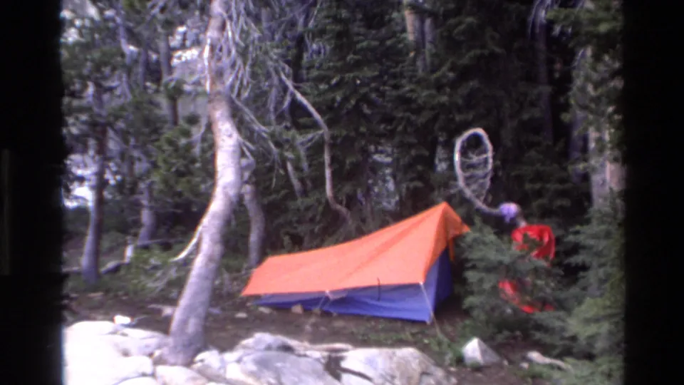 8mm home movie frame: orange-blue tent among conifers and boulders, red-clad figure near tent, circular hoop hanging from branch, California backcountry, c.1980.