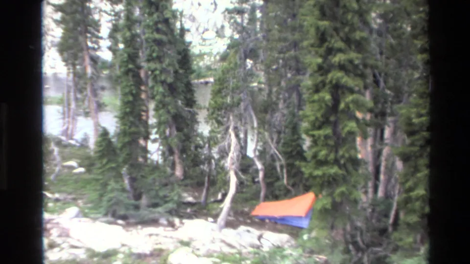 8mm frame revealing a wider forest view; orange tent at right margin, dense conifers, and a blue opening through the trees — possibly a lake or clearing — California backcountry, c.1980.