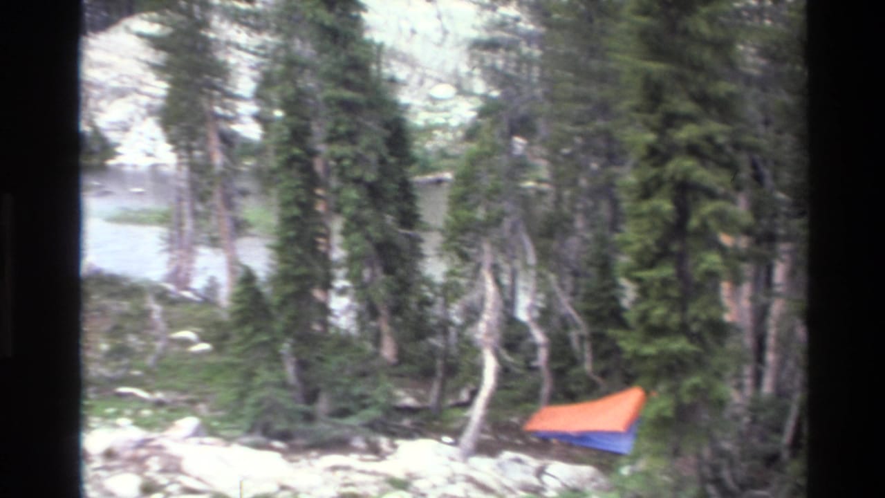 Orange and blue tent pitched on a rocky alpine lakeshore surrounded by sparse conifers; California backcountry, circa 1980, 8mm home movie footage.