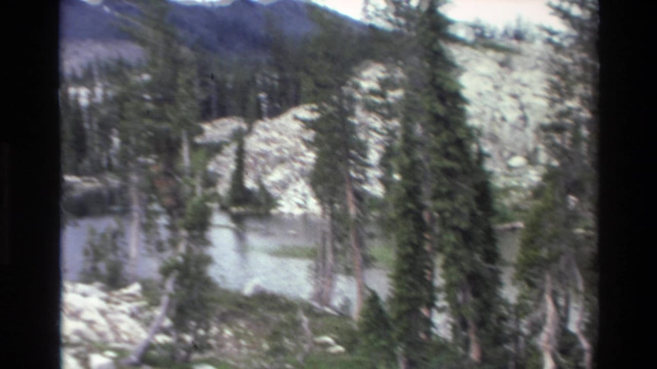 Mountain lake with rocky shore and sparse conifers; distant mountain ridge visible upper right; California backcountry, circa 1980, 8mm home movie.
