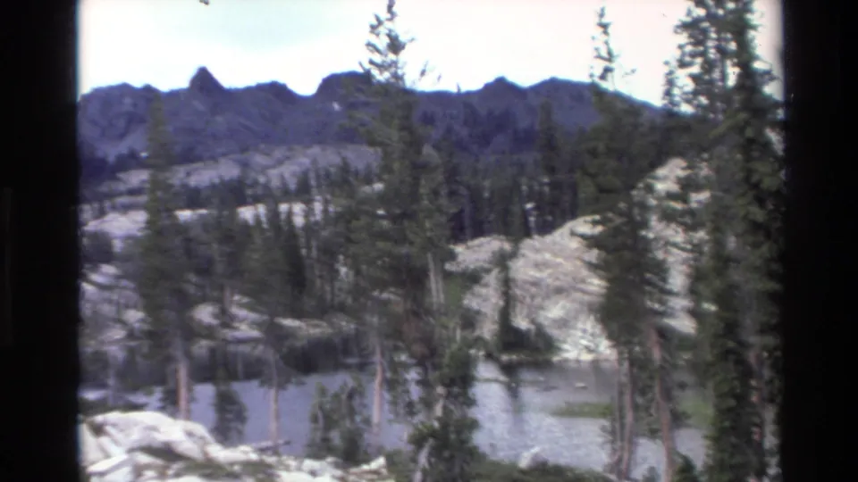 Dramatic jagged dark mountain ridge above calm alpine lake and rocky shore with sparse conifers; California backcountry, circa 1980, 8mm home movie footage.