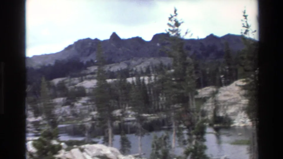 Wide pan frame of alpine lake, jagged mountain peaks, and rocky shoreline with sparse conifers; California backcountry, circa 1980, 8mm home movie footage.