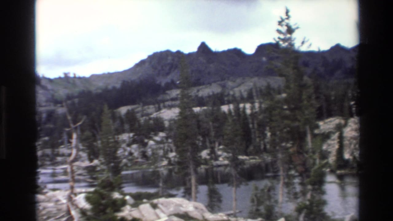 8mm home movie frame circa 1980 showing an alpine lake with granite boulder shoreline, dead snag trees, living pine forest, and rocky peaks in California mountain wilderness.