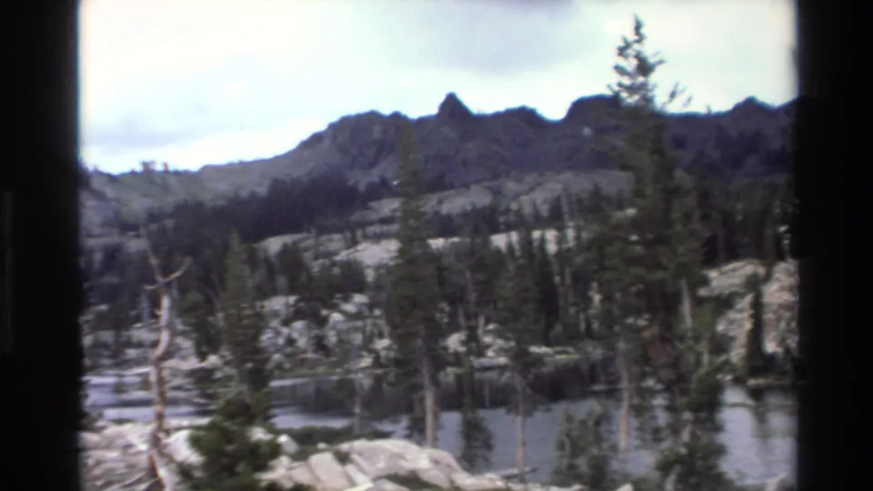 8mm home movie frame circa 1980 showing an alpine lake with granite boulder shoreline, dead snag trees, living pine forest, and rocky peaks in California mountain wilderness.