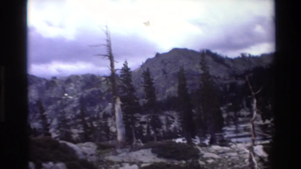8mm home movie frame circa 1980 completing a slow pan across rocky alpine wilderness with snag trees, conifers, and a dramatic overcast mountain ridge.