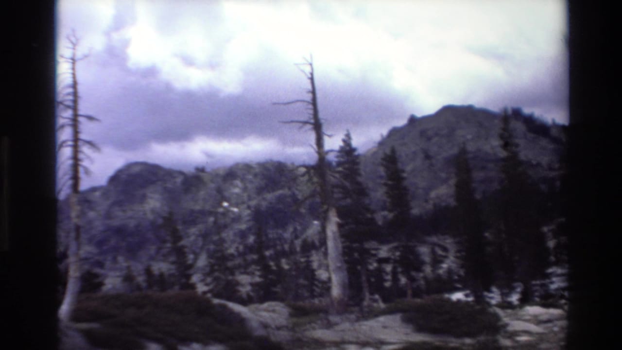 1980s 8mm home-movie frame: rugged mountain peak, skeletal snag trees, and dense conifers under a dramatic overcast sky in a California mountain setting.