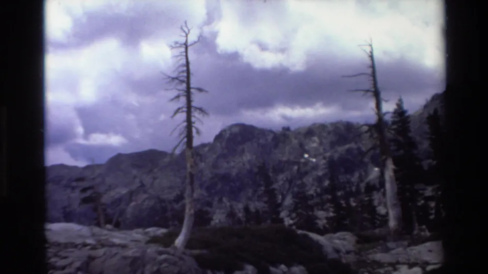 1980s 8mm home-movie frame: two skeletal dead trees in the foreground against a rocky California mountain peak and a heavy, billowing cloud sky.