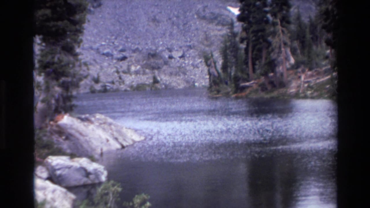 Alpine lake with rocky granite shoreline, conifers on far slope, and scree-covered mountain behind