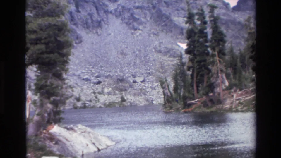 Wide shot of alpine mountain lake surrounded by scree slopes and evergreen trees, 8mm home movie footage labeled California, circa 1980