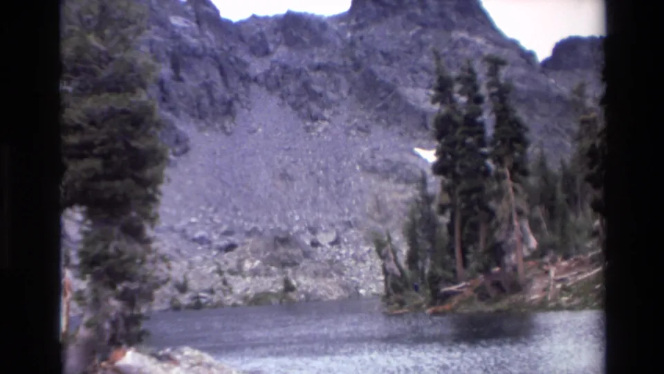 Alpine lake with snow-patched rocky slopes and evergreen tree line, 8mm home movie footage labeled California, circa 1980