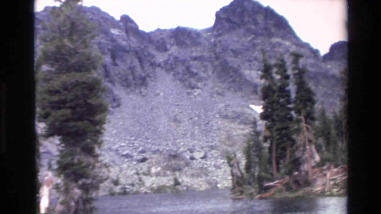 Alpine lake and talus slope beneath jagged dark peaks framed by conifers, California mountain wilderness circa 1980, scanned 8mm